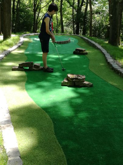 A person is playing mini-golf on a green course in the forest. Around him are trees and various obstacles made of stones.