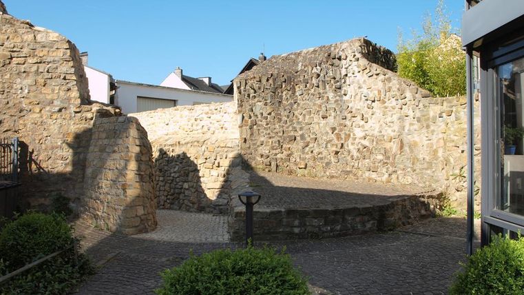An old stone wall in a quiet setting. The sky is clear and blue, surrounded by green plants.