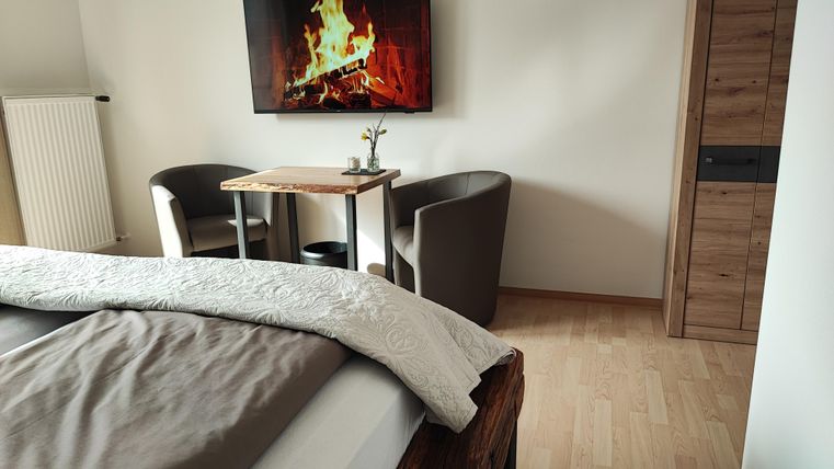 A cozy bedroom with a large bed and a television showing a fire. In the corner, there is a table and two comfortable chairs.