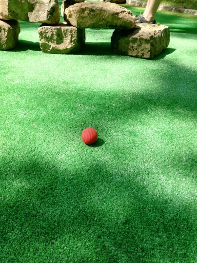 A red ball is lying on green artificial grass next to some stones. It is a quiet mini-golf setting.