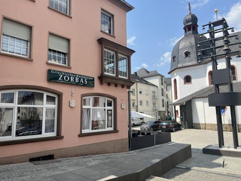 A street with a Greek restaurant named "Zorbas" and a church in the background. The sky is blue and there are some cars on the road.