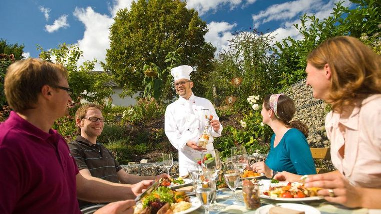 Un cuisinier sert un repas délicieux à une table en plein air. Cinq personnes apprécient le repas et l'ambiance agréable.