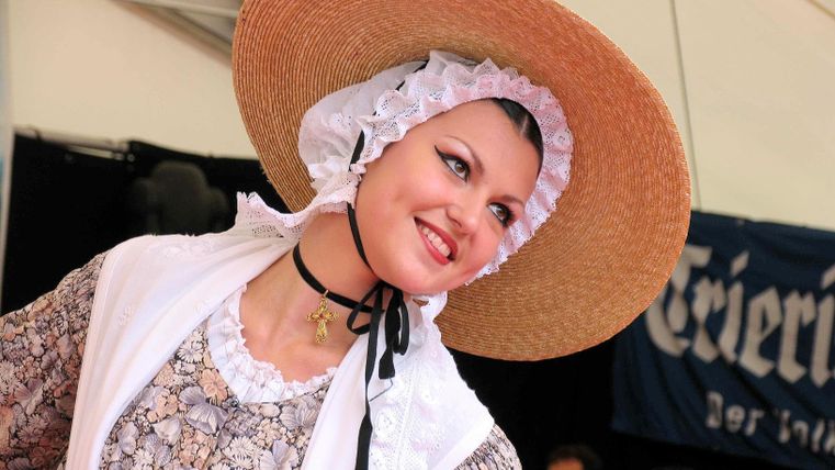 A woman in traditional costume with a large straw hat smiles happily. In the background, blurred people and a decorative stage are visible.