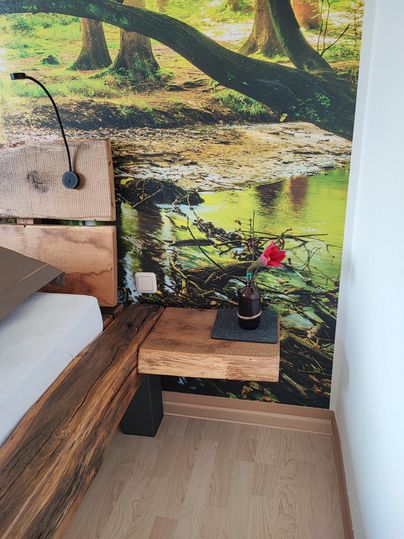 A cozy bedroom with a wooden bed and a nightstand. On the nightstand, there is a bottle and a red flower in front of a nature wallpaper.