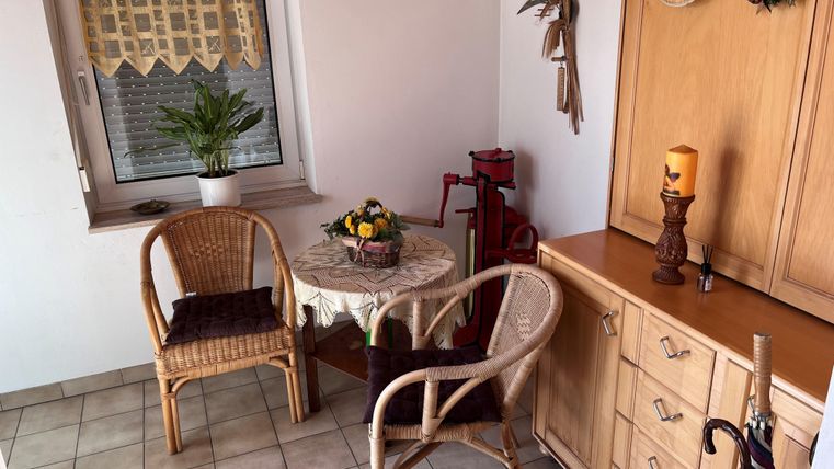 Een gezellige zitcorner met twee rotanstoelen en een tafel. Aan de muur hangen decoraties en bij het raam staat een plant.