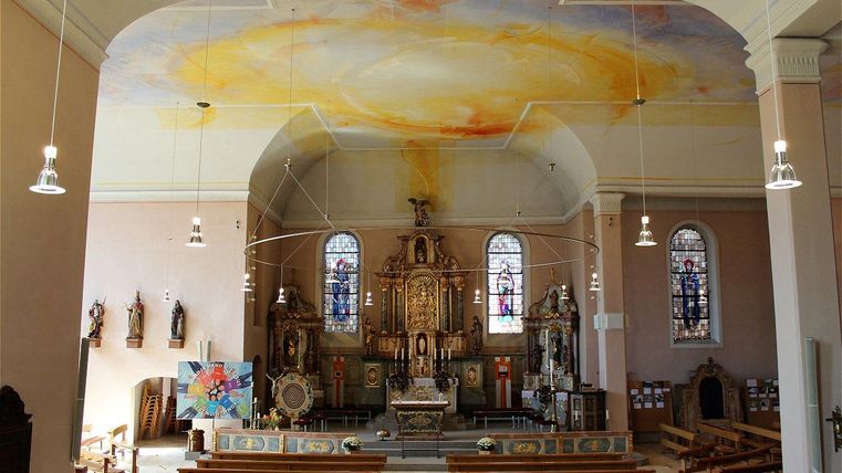 Eine serene Kirche mit einem beeindruckenden Deckenbemalung und bunten Fenstern. Die Bänke sind leer und der Altar hebt sich im Zentrum des Raumes hervor.