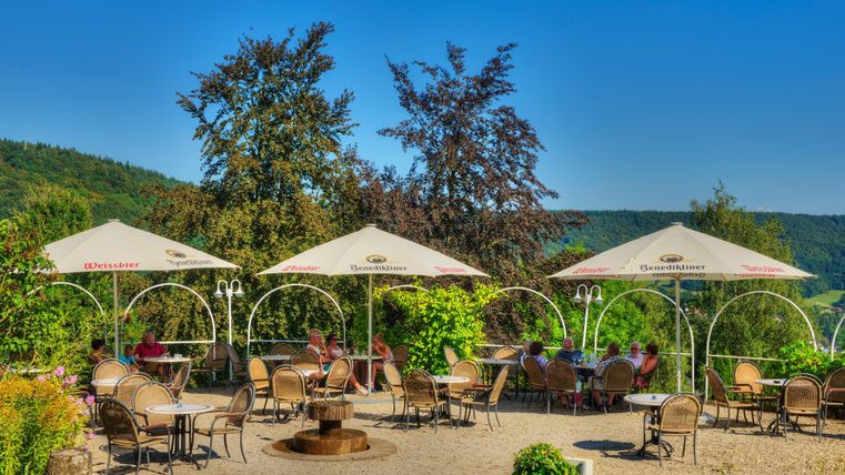 Ein gemütliches Café im Freien mit Schirmen und vielen Sitzgelegenheiten. Die Umgebung ist grün und von Bäumen umgeben.