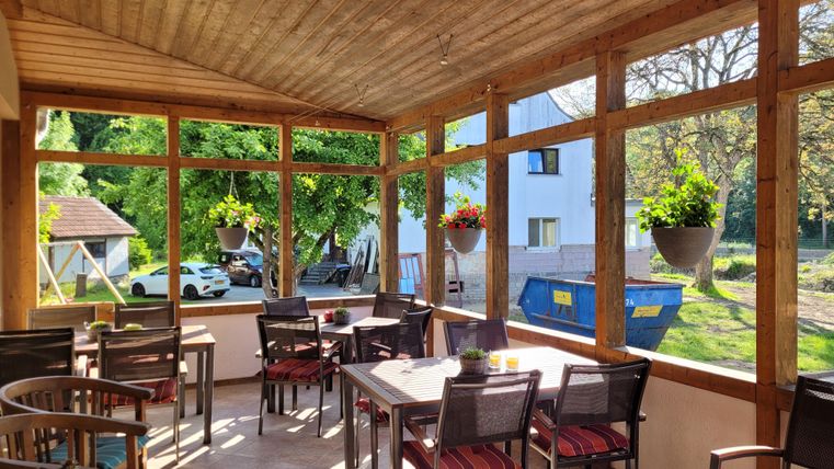 A beautiful conservatory with wooden walls and large windows. Several tables and chairs are set up, surrounded by plants and plenty of daylight.