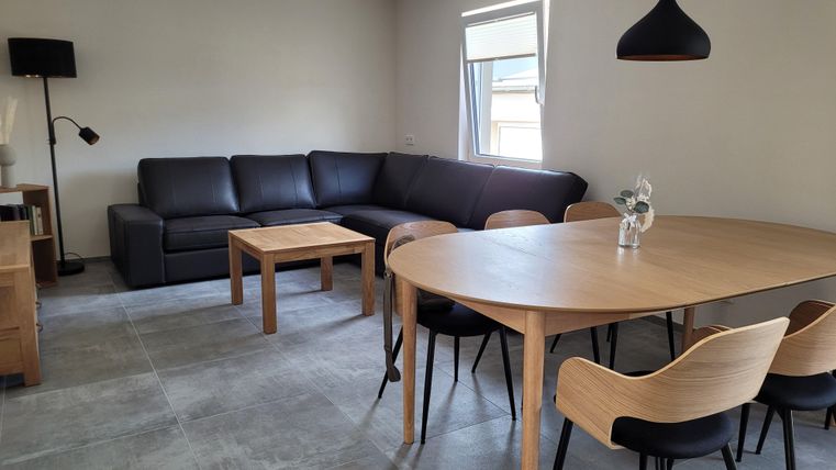 A modern living room with a large, comfortable sofa and a round dining table. The decor is simple and inviting, with a lamp and a window for natural light.