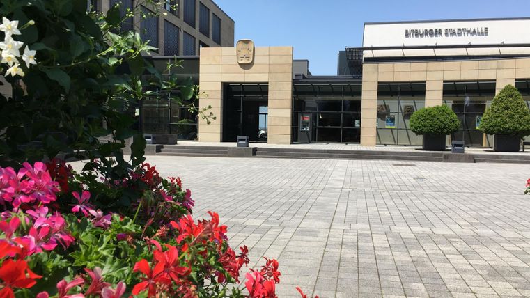 Ein modernes Gebäude mit einer Glasfassade und einem Eingangsbereich. Im Vordergrund blühen bunte Blumen und die Umgebung ist sonnig.