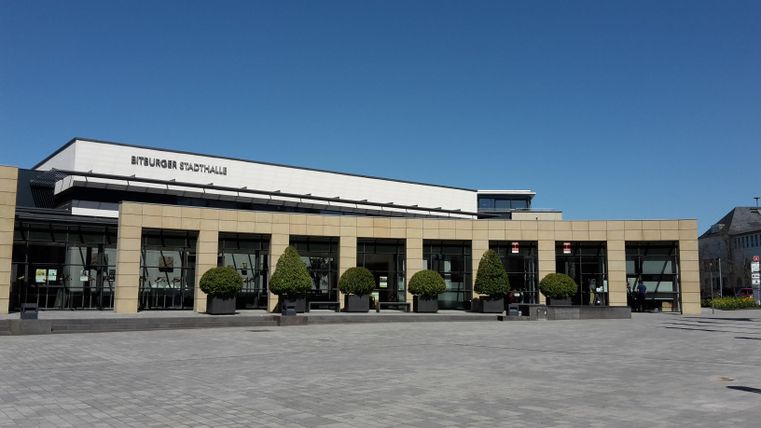 A modern building with large windows and a clear blue sky. In front of the building, there are evenly spaced trees and cobblestones.