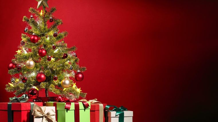 A festively decorated Christmas tree stands in front of a red wall. Beneath it are many lovingly wrapped gifts in various colors.