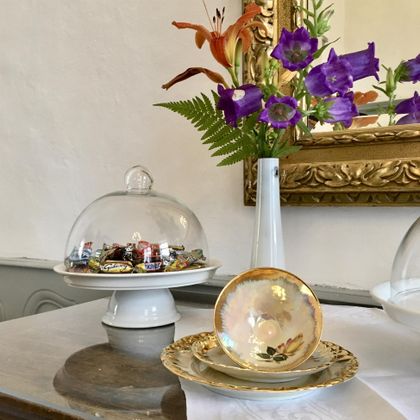 A beautiful table with a glass dome full of candies and an elegant porcelain cup. In the background, there are fresh flowers in a white vase and a golden frame on the wall.
