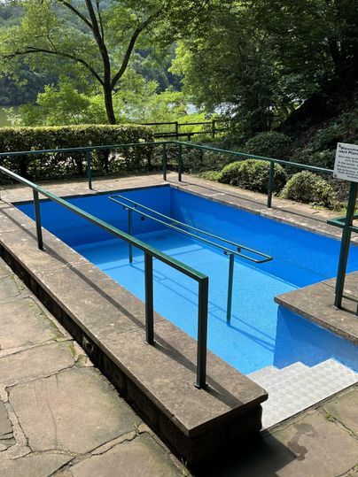 A small blue pool with railings in a green environment. In the background, trees and a calm lake are visible.