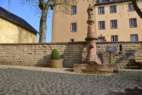 Eine historische Straße mit einem Steinbrunnen und einer alten Mauer. Im Hintergrund sind mehrere Fenster eines Gebäudes sichtbar.