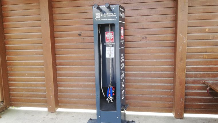 Eine Waschstation mit einem Schlauch und einem Wassertank steht vor einer Holzwand. Die Station ist für die Reinigung von Fahrrädern oder Ausrüstung gedacht.