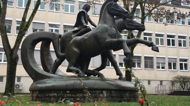 A bronze-like statue of a rider on a horse stands in a green area in front of a building. The statue depicts the rider in a calm posture with an raised sword.
