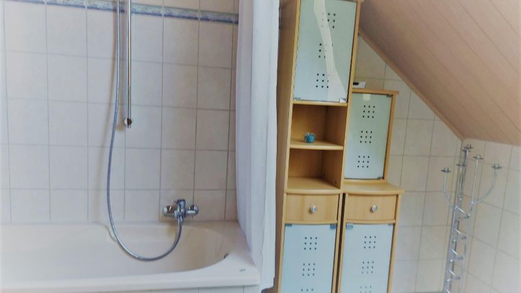 A bathroom with a bathtub and a shower installation. Next to the bathtub is a bright shelf with compartments for storage.
