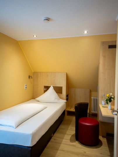 A cozy room with a single bed, a red stool, and a desk. The walls are painted in a warm shade of yellow, creating an inviting atmosphere.