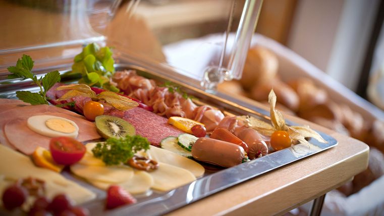 An attractively decorated platter with various types of sausage and cheese, fresh fruit and vegetables. In the background, rolls are visible that complement the breakfast buffet.