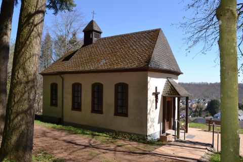 Eine kleine Kapelle umgeben von Bäumen und einem klaren Himmel. Der einladende Eingang führt zu einem ruhigen Außenbereich.