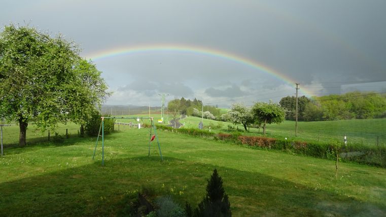 A green garden with a rainbow arching over the landscape. In the background, there are trees and meadows.