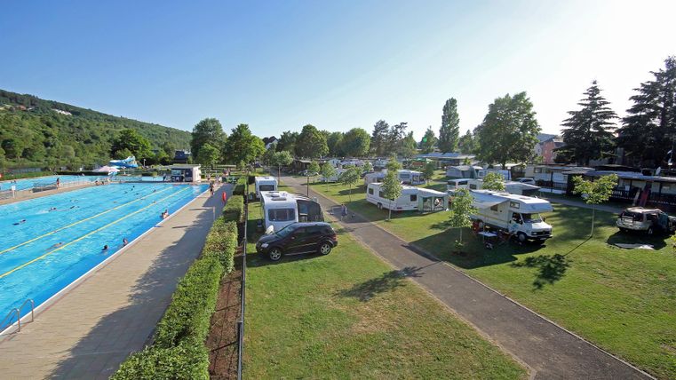 Ein Freibad mit blauen Wasserbecken und vielen Bäumen umgeben. Im Hintergrund sind Wohnmobile und ein grüner Campingplatz zu sehen.