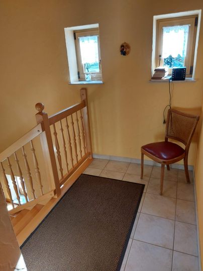 A bright entrance area with yellow walls and a staircase. A carpet lies on the floor and a chair stands next to a small window.