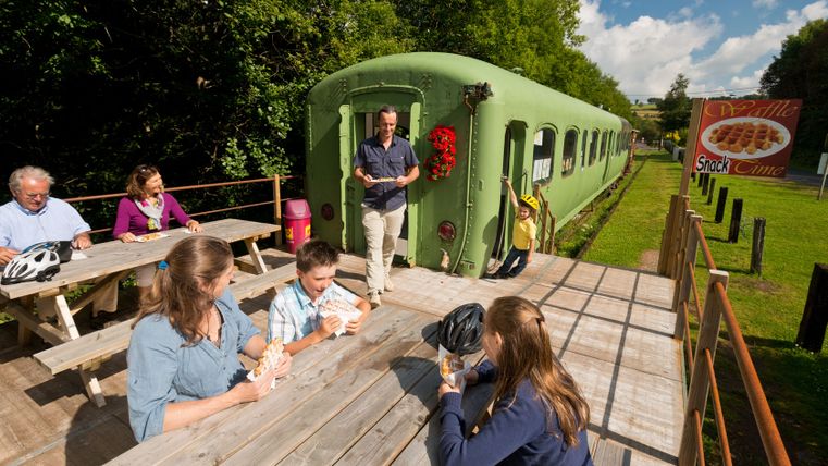Menschen essen Waffeln an Holztischen neben einem grünen Eisenbahnwaggon.