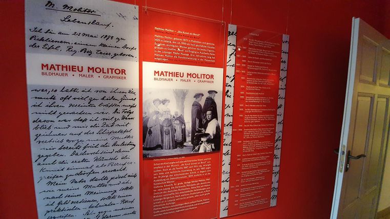 Een muur met informatie over Mathieu Molitor, inclusief handgeschreven teksten en foto's. De muur is rood geschilderd en toont verschillende tentoonstellingsstukken.