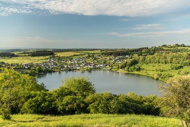 Ein malerischer Blick auf das Schalkenmehrener Maar in der Vulkaneifel bei sonnigem Wetter. Im Vordergrund dichter, grüner Laubwald, dahinter der kreisrunde Maarsee mit spiegelnder Wasseroberfläche. Umgeben ist der See von Wiesen, Feldern und dem kleinen Ort Schalkenmehren. Im Hintergrund erstreckt sich die hügelige Eifellandschaft unter einem blauen Himmel mit leichten Schleierwolken.
