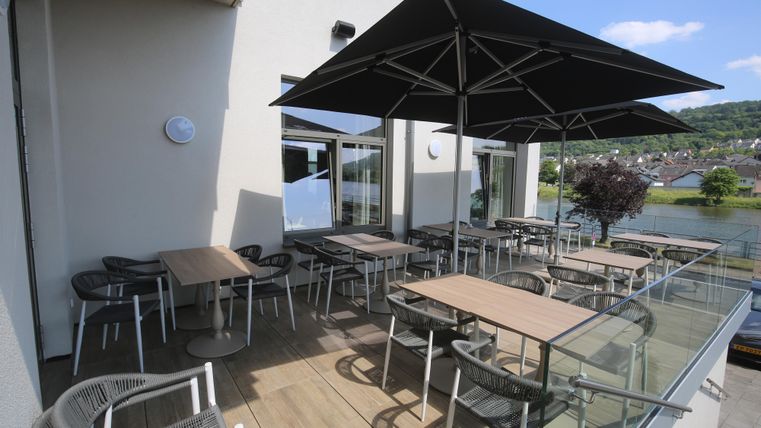 Een gezellige terras met tafels en stoelen is overdekt door een grote parasol. Op de achtergrond is een rustige waterlandschap te zien.