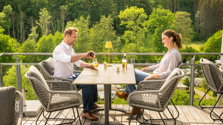 A couple is sitting at a table on a terrace enjoying drinks. In the background, green trees and a beautiful landscape can be seen.