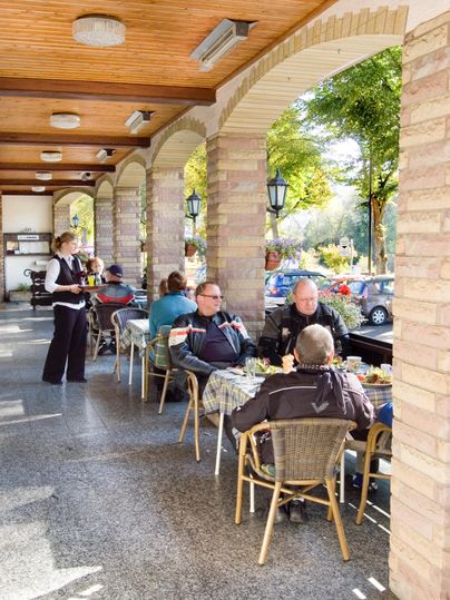 Eine gemütliche Terrasse mit mehreren Gästen, die Essen genießen. Eine Kellnerin serviert Speisen unter schattenspendenden Bäumen.