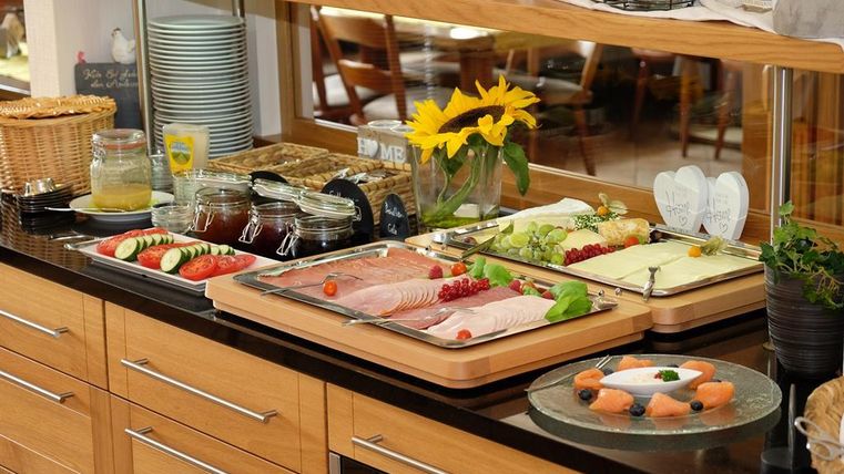 A rich breakfast buffet on a counter with cold cuts, fruit, and fresh vegetables. Various jams and drinks are also arranged.