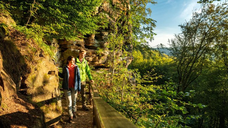 Zwei Wanderer gehen auf einem Weg entlang einer Felsenlandschaft. Die Umgebung ist grün und sonnig, mit Bäumen und Felsen im Hintergrund.