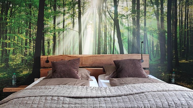 A cozy bedroom with a wooden bed and soft pillows. The wall features a beautiful forest with sunlight filtering through the trees.