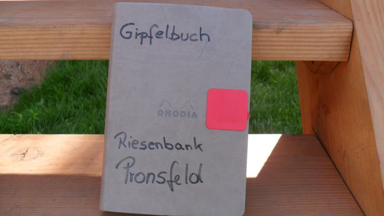 Un livre de sommet avec le titre "Riesenbank Pronsfeld" repose sur un banc en bois. Il a une couverture grise et une étiquette rouge.
