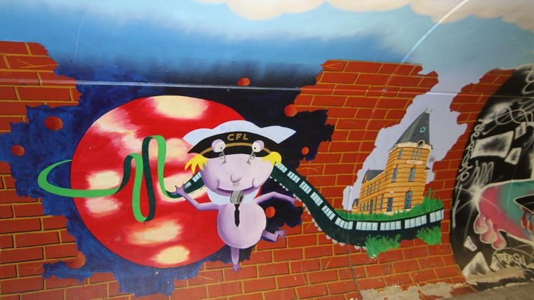 Een kleurrijke muurschildering toont een cartoonachtige figuur met een locomotief en een rode zon op de achtergrond. De scène is omgeven door een bakstenen muur.