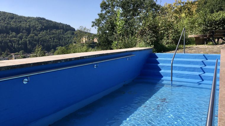A blue pool with a staircase, surrounded by trees and hills. The sky is clear and sunny.