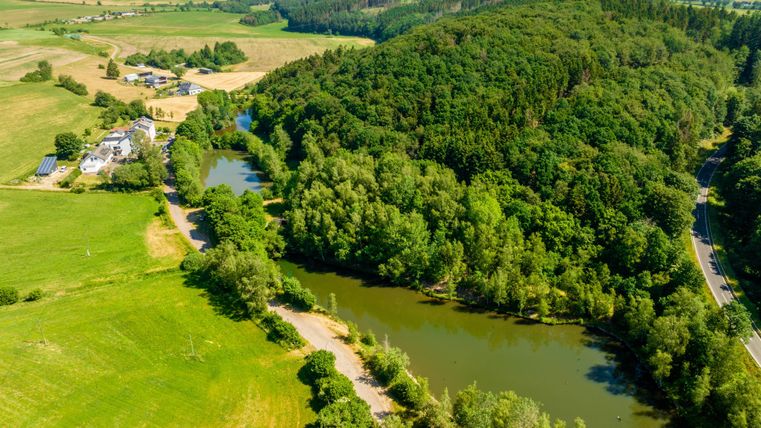 Un paysage pittoresque avec une rivière, entourée de verdure luxuriante et de collines boisées. À gauche, des champs et un bâtiment sont visibles.