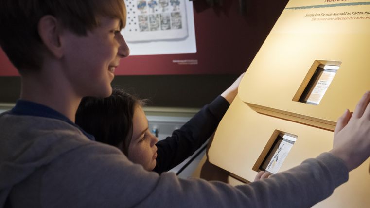 Deux enfants regardent avec intérêt un objet d'exposition interactif dans un musée. Ils touchent les écrans et semblent apprendre quelque chose.