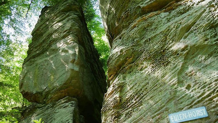 Eine beeindruckende Felsenformation umgeben von grünem Laub. Auf einem Felsen ist ein Schild mit der Aufschrift "Eulen-Horst" zu sehen.