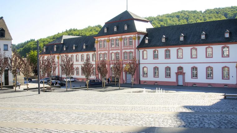 Un bâtiment historique avec une façade élégante se trouve sur une place pavée. À l'arrière-plan, on aperçoit des collines douces.