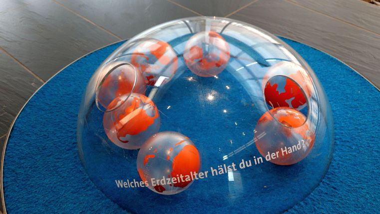 A glass dome with orange balls representing the Earth. Below it is the inscription "Which geological age are you holding in your hand?"
