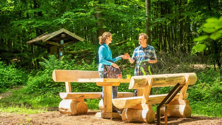 Zwei Personen stehen an einem Holztisch in einem Waldgebiet. Die Umgebung ist grün und freundlich mit Bänken aus Holz.