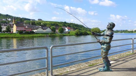 Eine Bronze-Statue eines Fischers steht am Ufer eines Flusses. Im Hintergrund sind malerische Häuser und grüne Hügel zu sehen.