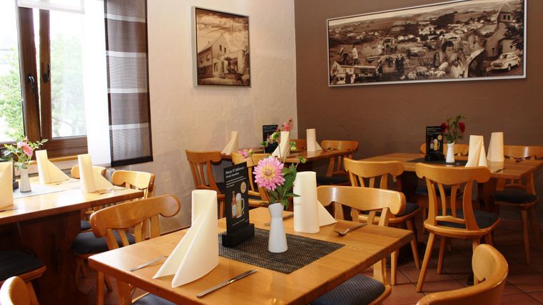 Ein einladendes Restaurant mit Holztischen und Stühlen. Vasen mit Blumen und frische Tischdecken schaffen eine angenehme Atmosphäre.