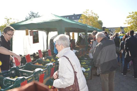 Een levendige weekmarkt met veel mensen die verse voedingsmiddelen kopen. Op de achtergrond staan groene verkoopstands en een parasol.