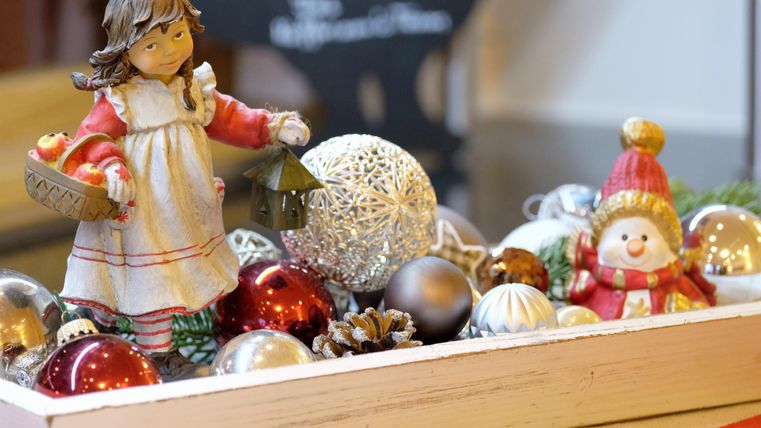 A festive decoration featuring a small figure of a girl holding a lantern. Surrounding her are colorful Christmas ornaments and a snowman figure.